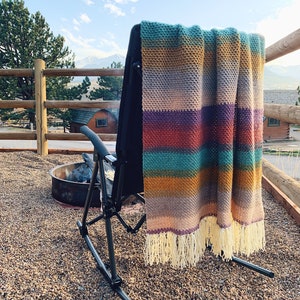 Pu&ograve; includere: Una coperta a righe in crochet con frange &egrave; appesa allo schienale di una sedia a dondolo nera su un patio in ghiaia. La coperta presenta una variet&agrave; di colori, tra cui il verde acqua, il viola, l'arancione e il giallo.