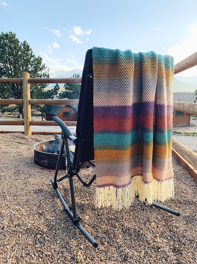Pu&ograve; includere: Una coperta a righe in crochet con frange &egrave; appesa allo schienale di una sedia a dondolo nera su un patio in ghiaia. La coperta presenta una variet&agrave; di colori, tra cui il verde acqua, il viola, l'arancione e il giallo.