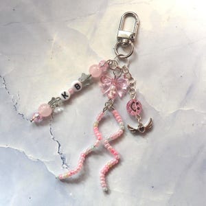 Peut inclure: Un porte-clés avec un mousqueton argenté attaché à une collection de perles roses, une breloque papillon rose translucide, une perle smiley rose et une breloque cœur ailé argenté. Un brin inclut des perles lettres blanches formant KB.