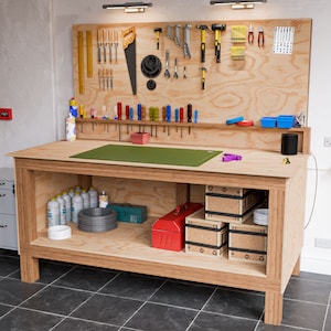 May include: A wooden workbench with a green work surface, shelves, and drawers. The workbench is filled with tools, including a saw, hammers, screwdrivers, and wrenches. There are also several containers of paint and other supplies.
