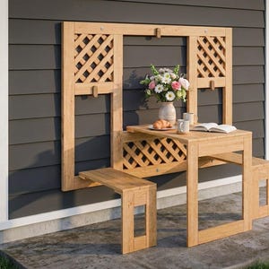 May include: A wooden outdoor folding table and bench set. The table and bench are made of light-colored wood and feature a lattice design. A vase of flowers, a book, and a croissant are on the table. The set is mounted on a dark gray wall.