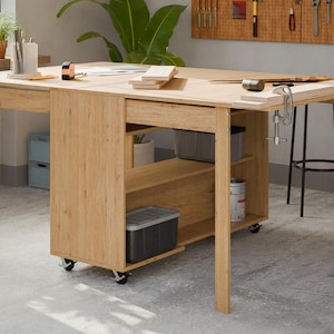 Puede incluir: Un banco de trabajo de madera de color claro con una extensión plegable, cajones y estantes. El banco de trabajo tiene un armario incorporado sobre ruedas y está equipado con abrazaderas. Las herramientas son visibles en un panel perforado en el fondo.