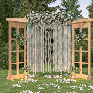 May include: A wooden wedding archway decorated with white floral garland and a macrame backdrop. The archway is set up outdoors in a grassy area with trees in the background.