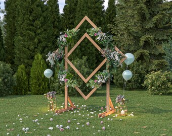 May include: A wooden diamond-shaped archway decorated with greenery and flowers, with two light blue balloons and lanterns on either side. The archway is set up in a grassy area with a forest background.
