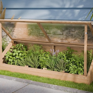 Puede incluir: Un invernadero de madera con una tapa transparente en ángulo, lleno de varias hierbas y plantas verdes. El marco está sobre un lecho de hierba verde, con una pared de ladrillo y revestimiento azul en el fondo.