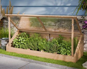 Könnte beinhalten: Ein hölzerner Frühbeetkasten mit einem transparenten, abgewinkelten Deckel, gefüllt mit verschiedenen grünen Kräutern und Pflanzen. Der Rahmen steht auf einem Bett aus grünem Gras, mit einer Backsteinmauer und blauen Seitenwänden im Hintergrund.