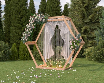 May include: A wooden hexagonal wedding arch decorated with white lace curtains and pink flowers. The arch is set up outdoors in a grassy area with trees in the background.