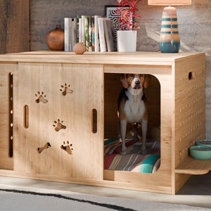 Op de afbeelding: Een houten hondenbench met een schuifdeur met uitsparingen in de vorm van pootafdrukken. Een beagle staat binnen op een gestreepte mat. De bench heeft een plank met twee turquoise kommen. De bench is van licht hout.