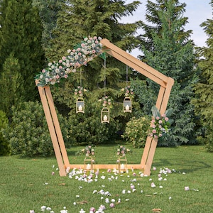 May include: A wooden wedding arch decorated with pink flowers and hanging lanterns. The arch is set up in a grassy area with trees in the background.