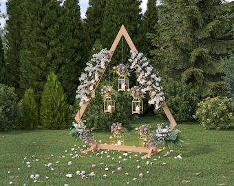May include: A wooden triangular archway decorated with white and pink flowers and hanging lanterns. The archway is set up outdoors in a grassy area with green trees in the background.