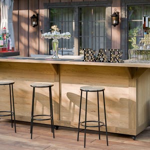 Puede incluir: Bar al aire libre con acabado de madera clara, con grifo, taburetes y letrero luminoso "BAR". El bar está instalado en una terraza de madera con un sofá gris y una parrilla en el fondo. Se exhiben varias botellas y vasos.