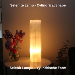 Könnte beinhalten: Eine weiße Selenitlampe in zylindrischer Form, die leuchtet und einen warmen Schein abgibt. Die Lampe steht auf einem weißen Tisch mit einer Blumenvase im Hintergrund. Der Text "Selenite Lamp - Cylindrical Shape" befindet sich oben im Bild und "Selenit Lampe - zylindrische Form" unten im Bild.
