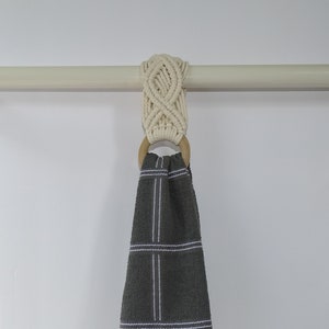 May include: A white macrame towel ring with a wooden ring hanging on a white rod. A gray kitchen towel with white stripes is hanging on the ring.