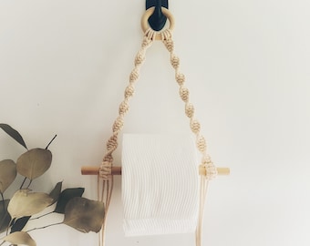 May include: A macrame toilet paper holder with a wooden dowel and a natural rope design. The holder is hanging from a black hook on a white wall.