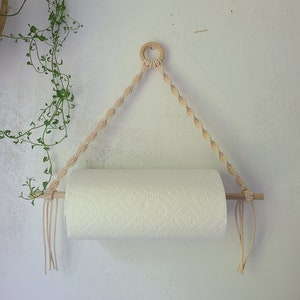 May include: A white paper towel roll hanging on a wooden dowel rod with a macrame rope holder. The rope is beige and has a wooden ring at the top.