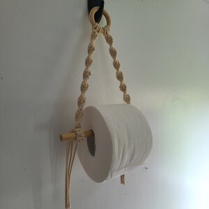 Puede incluir: Un rollo de papel higi&eacute;nico blanco colgado de un soporte de cuerda de macram&eacute; con una clavija de madera y un gancho negro.