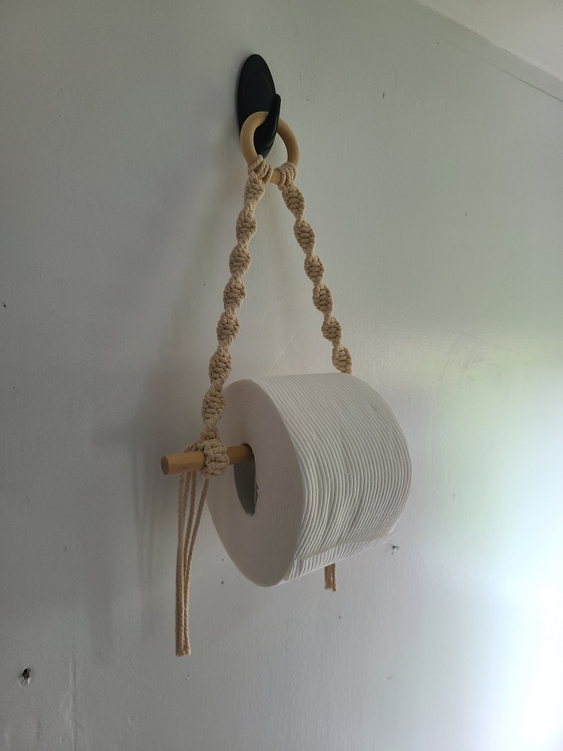 Puede incluir: Un rollo de papel higi&eacute;nico blanco colgado de un soporte de cuerda de macram&eacute; con una clavija de madera y un gancho negro.