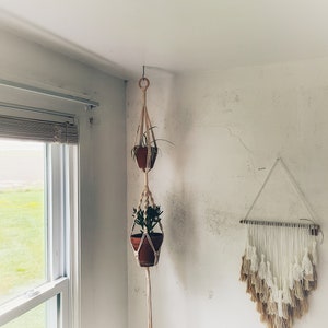 May include: Two potted plants hanging from a macrame plant hanger in a corner of a room. The plants are in terracotta pots and the macrame is cream colored. A white and beige macrame wall hanging is on the wall to the right.