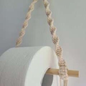 Puede incluir: Un soporte de papel higi&eacute;nico de madera con un dise&ntilde;o de cuerda natural. El soporte est&aacute; colgado de un anillo de madera y tiene un rollo de papel higi&eacute;nico blanco encima.