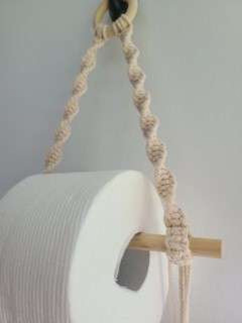 Puede incluir: Un soporte de papel higi&eacute;nico de madera con un dise&ntilde;o de cuerda natural. El soporte est&aacute; colgado de un anillo de madera y tiene un rollo de papel higi&eacute;nico blanco encima.