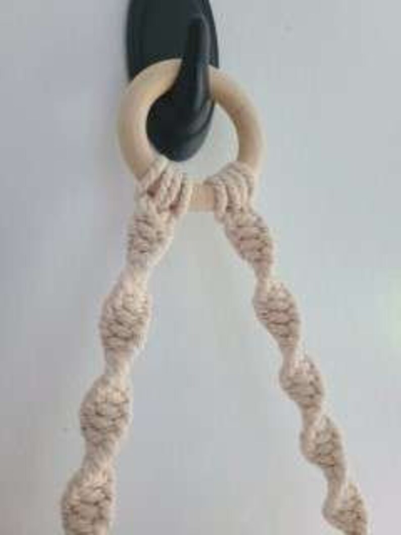 Puede incluir: Primer plano de un macram&eacute; para colgar plantas con un anillo de madera. El macram&eacute; est&aacute; hecho de cuerda de algod&oacute;n natural y tiene un dise&ntilde;o en espiral. El macram&eacute; est&aacute; sujeto a un gancho negro.