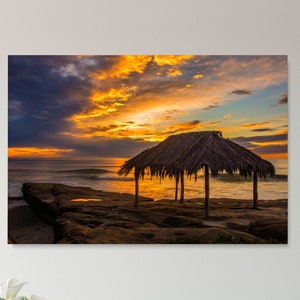 Puede incluir: Una cabaña con techo de paja se alza sobre una playa rocosa al atardecer. El cielo es una mezcla vibrante de nubes naranja, amarillas y rosadas. El océano está tranquilo y el agua es de un azul profundo.
