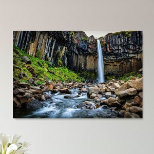 May include: A framed print of a waterfall cascading down a cliff face, surrounded by dark rock formations and green vegetation. The water flows into a rocky stream. The print is displayed above a white table with wooden chairs and a vase of white flowers.