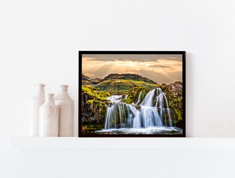 May include: A framed photograph of a waterfall cascading down a mossy hillside. The sun shines through the clouds, creating a dramatic light effect. The image is in a black frame.