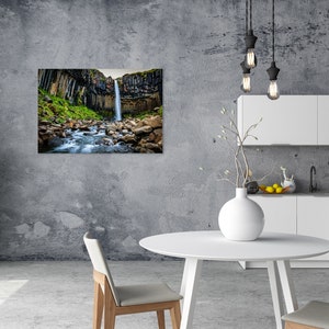 May include: A framed landscape print of a waterfall cascading down a cliff face, surrounded by green vegetation and rocks. The print is displayed on a gray wall in a modern kitchen setting with white cabinets and a round table.