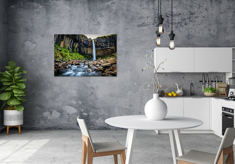 May include: A framed landscape print of a waterfall cascading down a cliff face, surrounded by green vegetation and rocks. The print is displayed on a gray wall in a modern kitchen setting with white cabinets and a round table.