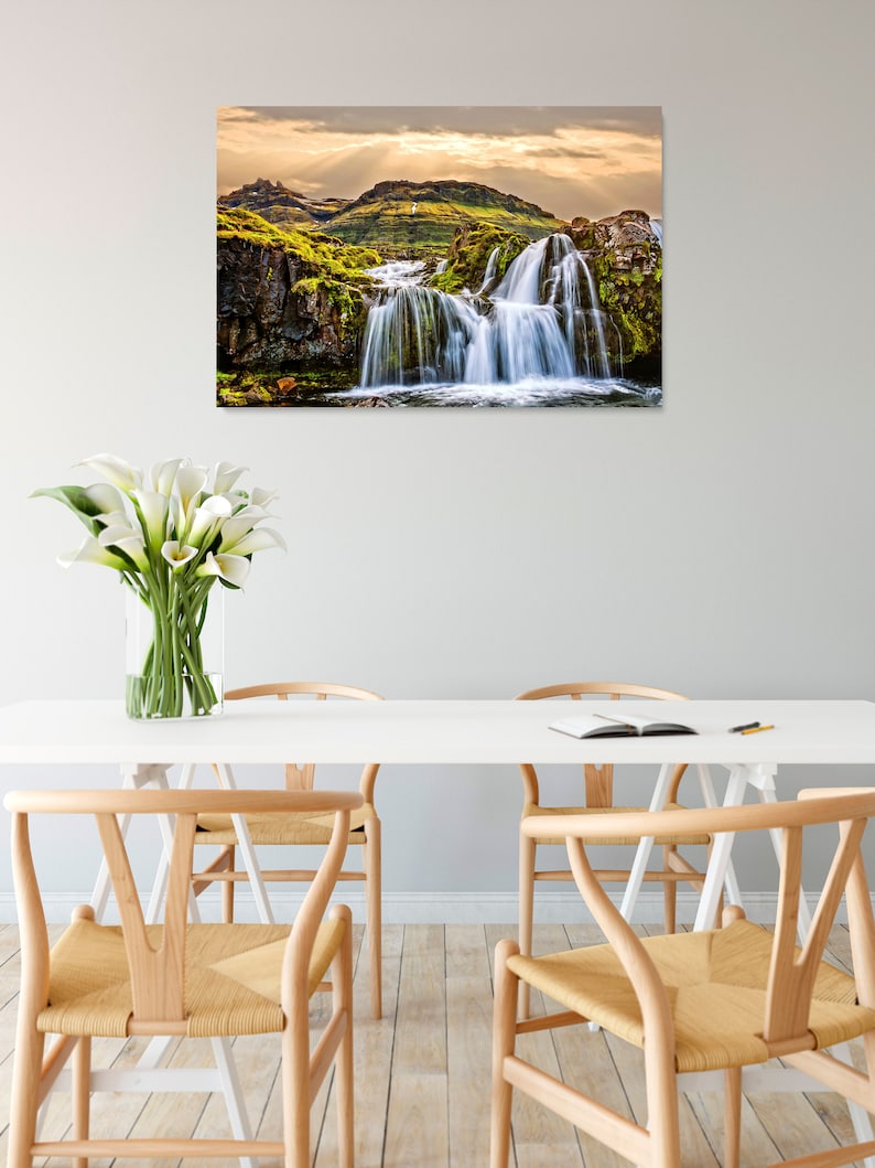 May include: A scenic photograph of a waterfall cascading over moss-covered rocks in a lush green landscape. The water is flowing over the rocks and creating a misty spray. The sky is a soft blue with white clouds.