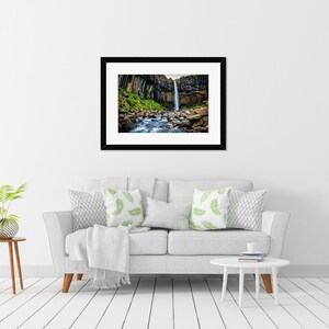 May include: A framed photograph of a waterfall cascading down a cliff face. The black framed print hangs above a white sofa with decorative pillows. A small side table with a plant and a white coffee table complete the room.