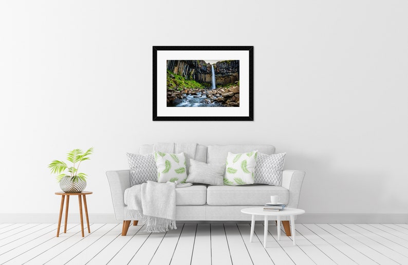 May include: A framed photograph of a waterfall cascading down a cliff face. The black framed print hangs above a white sofa with decorative pillows. A small side table with a plant and a white coffee table complete the room.