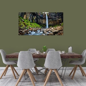 May include: A framed landscape print of a waterfall cascading down a cliff face. The waterfall is surrounded by dark, columnar rock formations and lush green vegetation. A stream flows through the foreground, with large rocks. The print is displayed above a dining table.