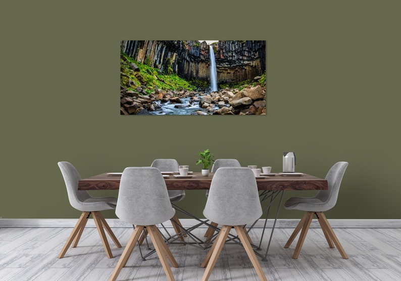 May include: A framed landscape print of a waterfall cascading down a cliff face. The waterfall is surrounded by dark, columnar rock formations and lush green vegetation. A stream flows through the foreground, with large rocks. The print is displayed above a dining table.
