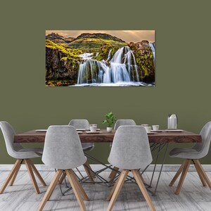 May include: A photograph of a waterfall cascading down a rocky cliff face covered in green moss. The waterfall is surrounded by lush greenery and a blue sky.
