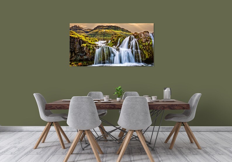 May include: A photograph of a waterfall cascading down a rocky cliff face covered in green moss. The waterfall is surrounded by lush greenery and a blue sky.
