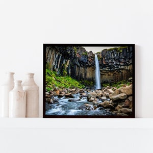 May include: A framed photograph of a waterfall cascading down a cliff face. The waterfall is surrounded by dark, columnar rock formations and lush green vegetation. A stream flows through the foreground, with rocks scattered throughout.