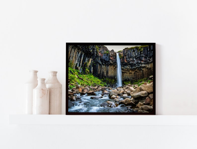 May include: A framed photograph of a waterfall cascading down a cliff face. The waterfall is surrounded by dark, columnar rock formations and lush green vegetation. A stream flows through the foreground, with rocks scattered throughout.