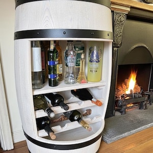 Peut inclure: Un meuble à vin en forme de tonneau en bois blanc avec des bandes métalliques noires. L'armoire possède des étagères pour les bouteilles et les verres à vin. Plusieurs bouteilles de vin et de liqueur sont visibles à l'intérieur. Le meuble est près d'une cheminée.