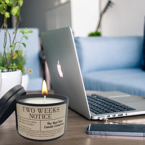 May include: A black candle with a brown label that reads "Two Weeks Notice" and "The Bad Idea Candle Company". The candle is lit and sitting on a wooden table with a laptop, a mobile phone, and potted plants in the background.