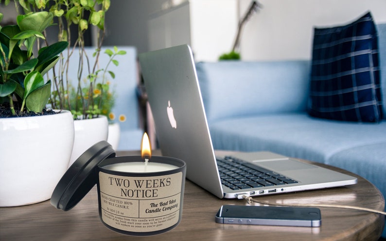 May include: A black candle with a brown label that reads "Two Weeks Notice" and "The Bad Idea Candle Company". The candle is lit and sitting on a wooden table with a laptop, a mobile phone, and potted plants in the background.