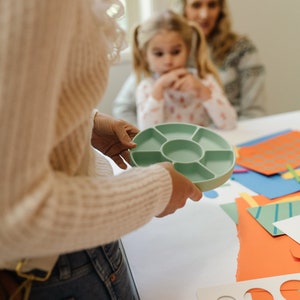 May include: A light green plastic tray with seven compartments, held by a person's hands. The tray is on a white table with colorful paper and craft supplies.