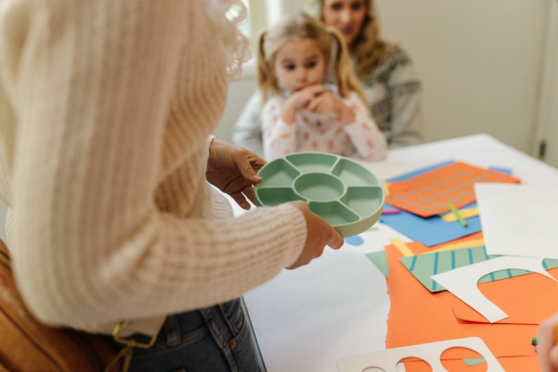May include: A light green plastic tray with seven compartments, held by a person's hands. The tray is on a white table with colorful paper and craft supplies.