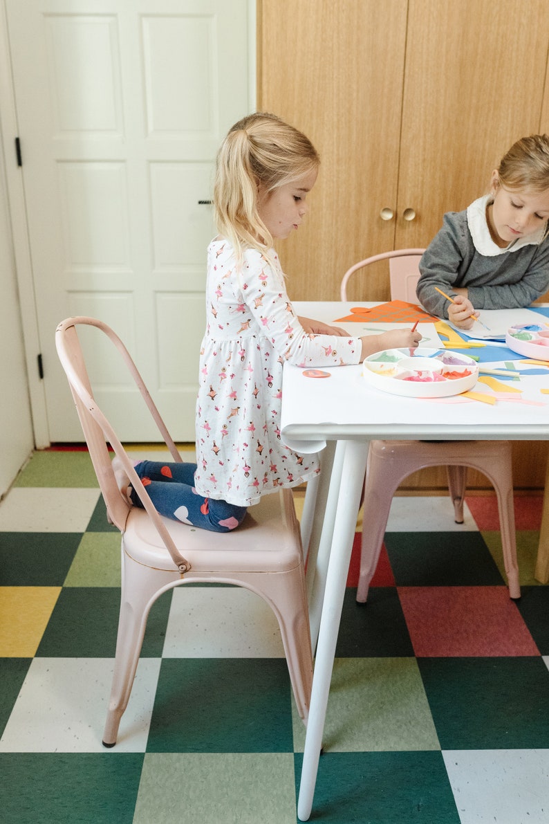 May include: Two children sit at a white table with pink chairs, creating art projects with colorful paper and paint. The table has a white paper tablecloth and a paint palette with colorful paints.