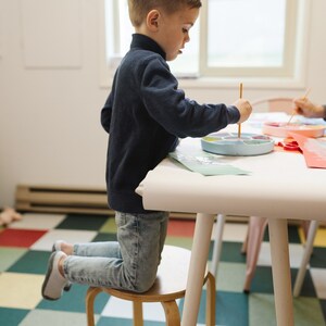 May include: A young child kneels on a wooden stool while painting with a paintbrush and a colorful paint palette. The child is wearing a blue sweater and jeans. The child is sitting at a white table with a checkered floor.