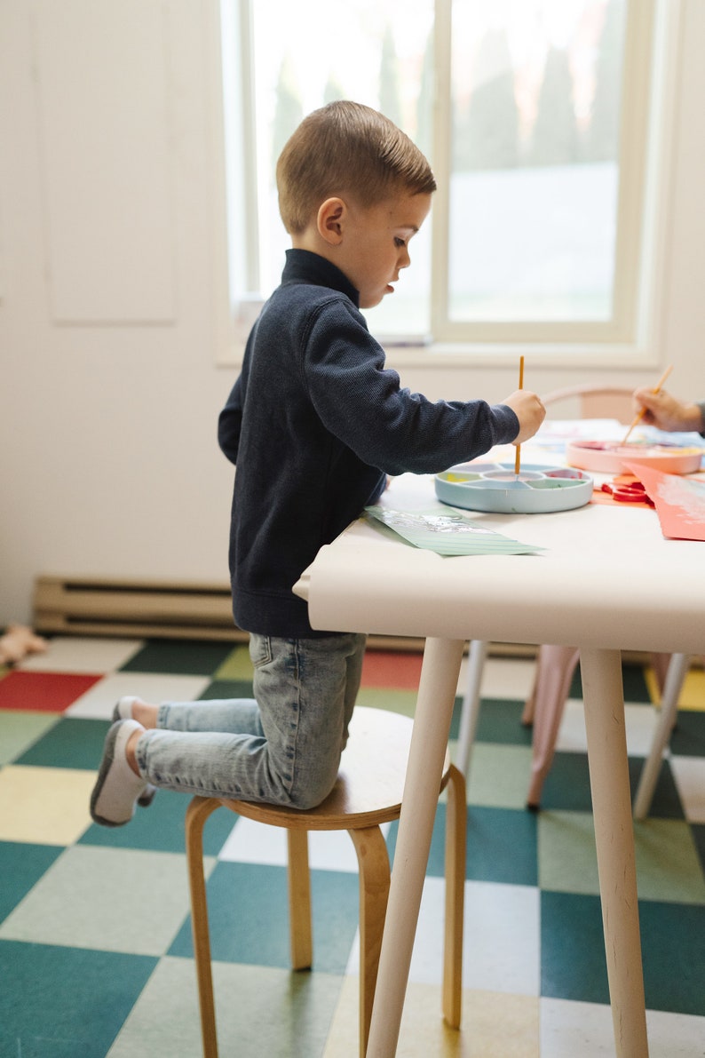 May include: A young child kneels on a wooden stool while painting with a paintbrush and a colorful paint palette. The child is wearing a blue sweater and jeans. The child is sitting at a white table with a checkered floor.