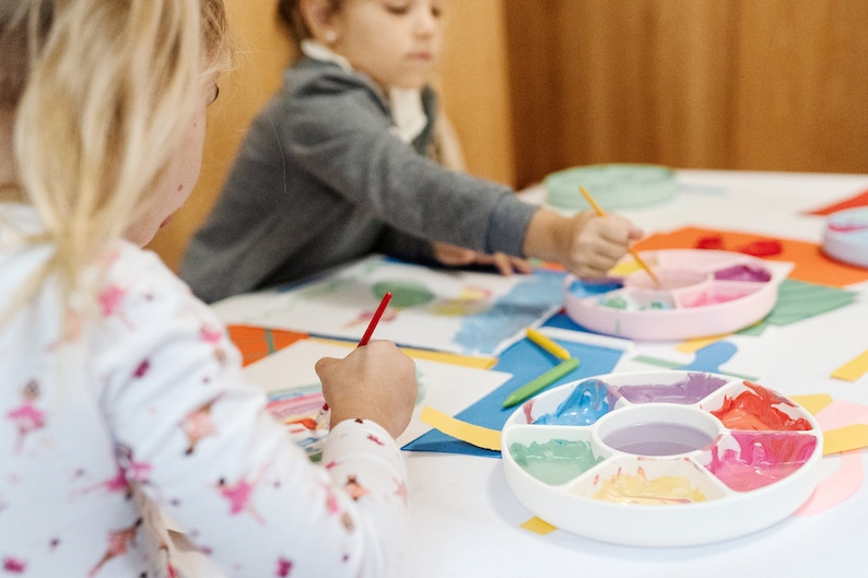 May include: Two children are painting with colorful paints on a white table. One child is using a red paintbrush and the other is using a yellow paintbrush. There are two paint palettes with different colors of paint.