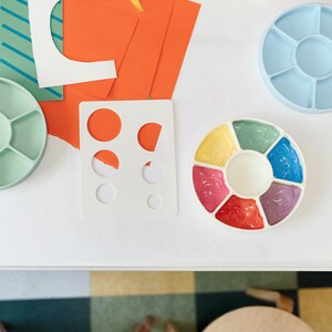 May include: A white plastic paint palette with six circular wells filled with different colored paints. The paints are yellow, green, blue, pink, red, and purple. The palette is on a white table with other art supplies.