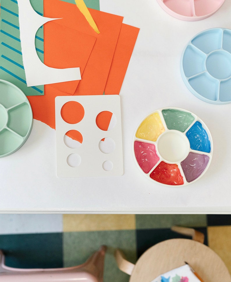 May include: A white plastic paint palette with six circular wells filled with different colored paints. The paints are yellow, green, blue, pink, red, and purple. The palette is on a white table with other art supplies.